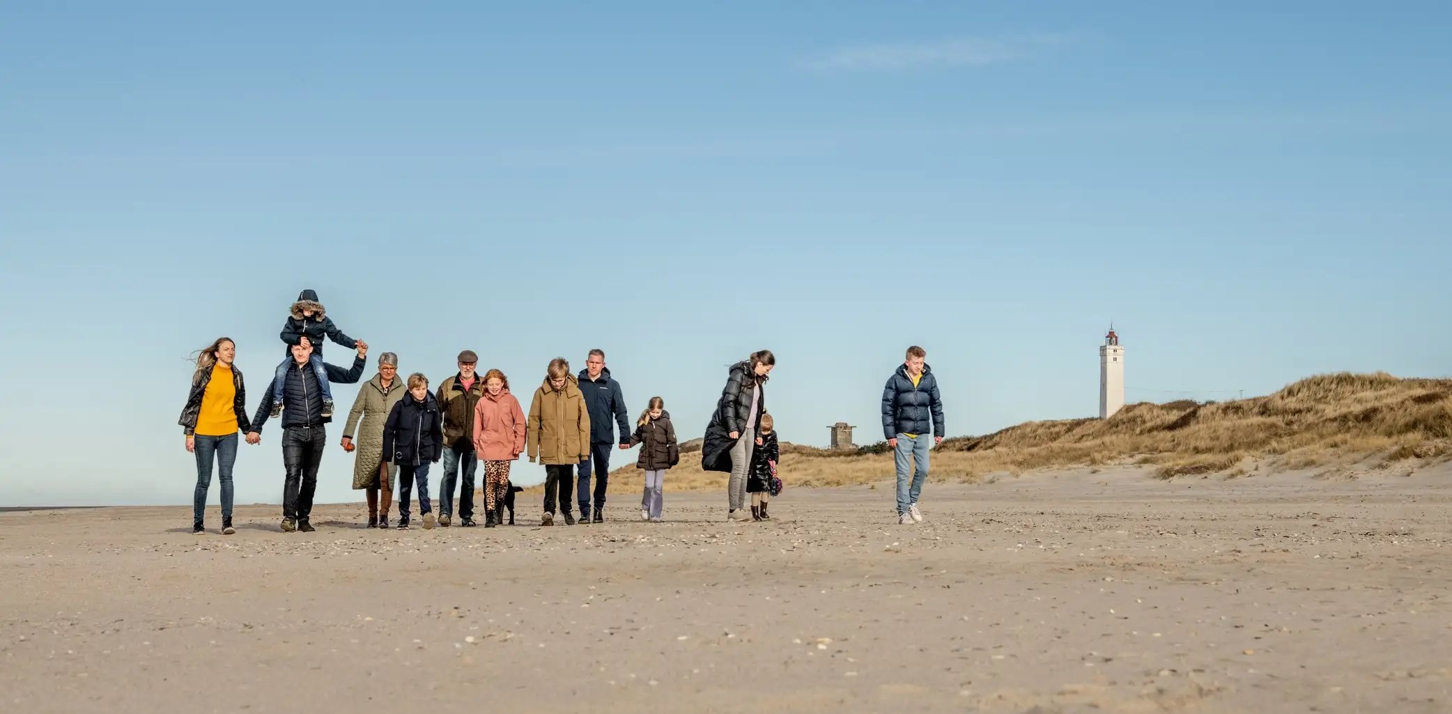 En familie går på en strand med klitter i baggrunden, der repræsenterer områdets naturskønne kystlinje.